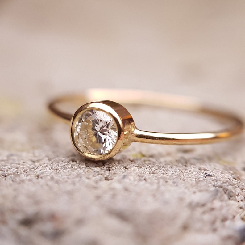May include: A delicate gold ring with a round, clear gemstone set in a bezel setting. The ring's band is thin and polished, and the gemstone sparkles against the neutral background. The ring is a piece of jewelry.