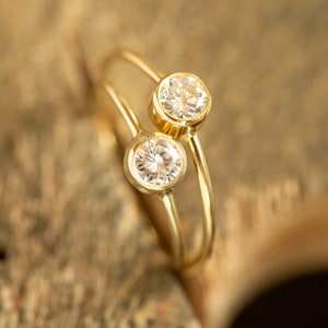 May include: A gold-colored ring with two clear, round gemstones. The gemstones are set in a bezel setting, and the band curves around the finger. The ring is displayed on a wooden surface.
