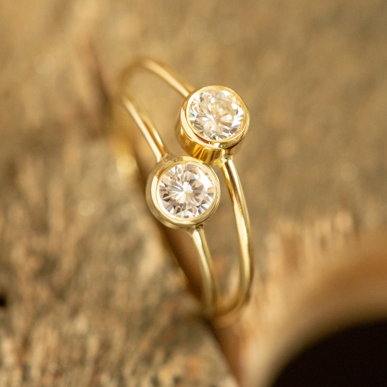 May include: A gold-colored ring with two clear, round gemstones. The gemstones are set in a bezel setting, and the band curves around the finger. The ring is displayed on a wooden surface.