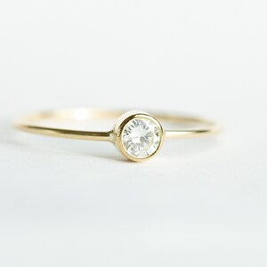 May include: A delicate gold ring with a round, clear gemstone set in a bezel. The ring has a thin band and a simple, elegant design. The gemstone is the focal point, catching the light and adding sparkle.