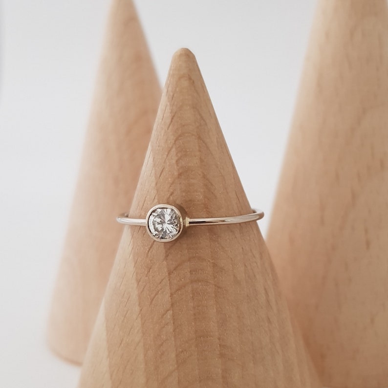 May include: A delicate silver ring with a round, clear gemstone set in a bezel. The ring is displayed on a wooden cone against a white background. The gemstone appears to be approximately 0.25 inches in diameter.
