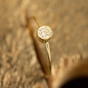 May include: A delicate gold ring featuring a single, round, clear gemstone. The gemstone is set in a bezel setting, and the band is thin and polished. The ring is displayed against a blurred, textured, brown background.