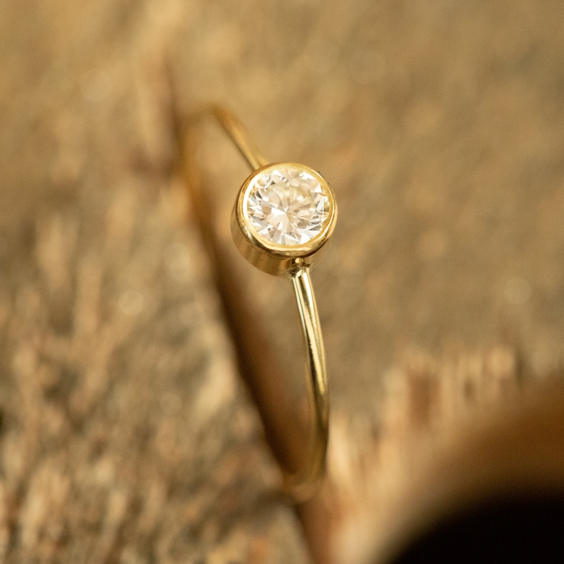 May include: A delicate gold ring featuring a single, round, clear gemstone. The gemstone is set in a bezel setting, and the band is thin and polished. The ring is displayed against a blurred, textured, brown background.