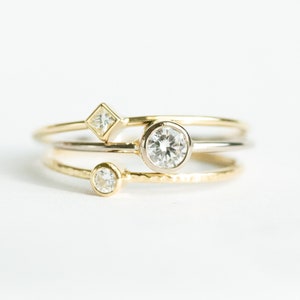 May include: Three delicate rings with clear gemstones. One ring features a round gemstone, another a square gemstone, and the third a smaller round gemstone. The rings are gold and silver in color, set against a white background.