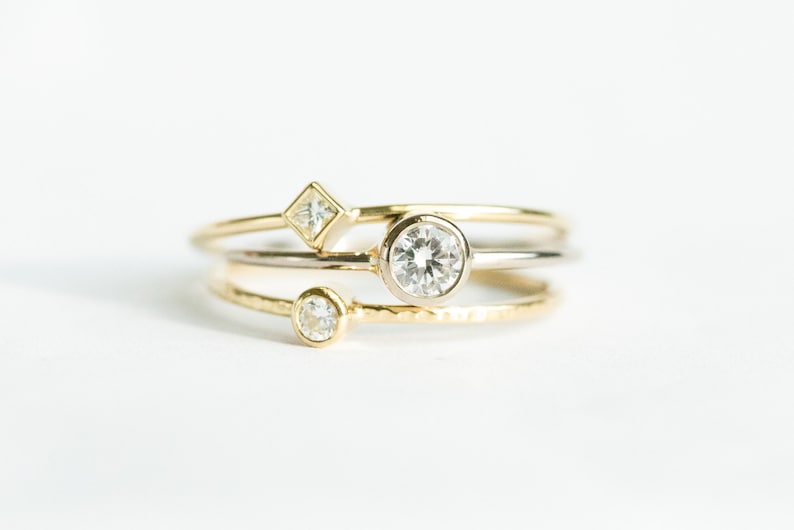May include: Three delicate rings with clear gemstones. One ring features a round gemstone, another a square gemstone, and the third a smaller round gemstone. The rings are gold and silver in color, set against a white background.