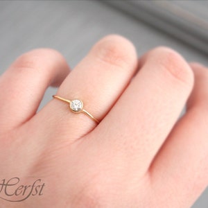May include: A delicate gold ring with a small, clear gemstone set in a bezel. The ring is on a slender band and is shown on a person's finger. The ring is a simple, elegant piece of jewelry.
