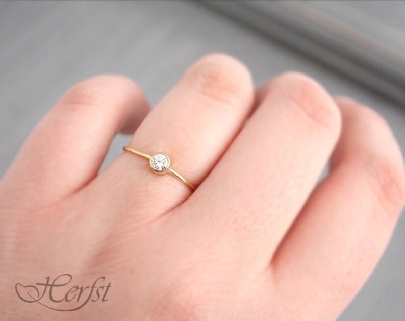 May include: A delicate gold ring with a small, clear gemstone set in a bezel. The ring is on a slender band and is shown on a person's finger. The ring is a simple, elegant piece of jewelry.
