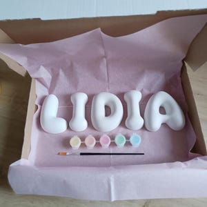 Op de afbeelding: Een set van vijf witte, afgeronde, driedimensionale letters die de naam LIDIA vormen, vergezeld van een kleine strip met vijf pastelkleurige verfpotjes en een dun penseel, alles gerangschikt in een kartonnen doos bekleed met lichtroze vloeipapier.