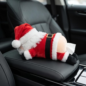Puede incluir: Un dispensador de pañuelos de peluche con forma de Papá Noel tumbado boca abajo. La figura lleva un traje rojo y blanco con un cinturón negro y una hebilla dorada. Un pañuelo blanco sale de la parte trasera del peluche, situado sobre una consola central de cuero negro.