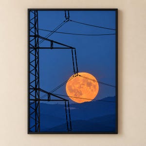 May include: A framed wall art print featuring a silhouette of a tall metal electrical transmission tower against a deep blue twilight sky. A large, bright orange full moon sits low on the horizon behind the dark, layered mountain ridges.