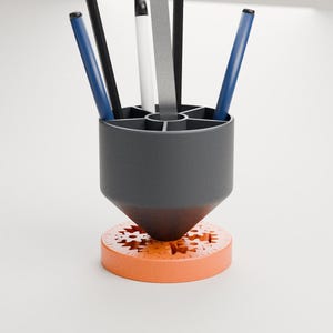 May include: A dark grey cylindrical pencil holder with six internal compartments, containing two blue pens, two black pens, one white pen, and one silver ruler. The holder rests on a circular orange base featuring visible internal gear mechanisms.