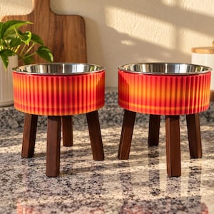 May include: Two elevated pet bowls featuring stainless steel inserts and ribbed, gradient-colored orange and red exterior bases. Each bowl is supported by three dark brown wooden legs, providing a raised feeding station for pets.
