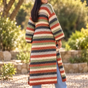 Könnte beinhalten: Eine lange, vorne offene Häkelstrickjacke mit horizontalen Streifen in Rostorange, Olivgrün, Creme und Anthrazit. Das Kleidungsstück hat lange, voluminöse Ärmel mit gerippten Bündchen und einen geraden, bodenlangen Saum.