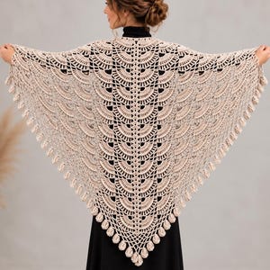 May include: A triangular crochet shawl in a light beige color featuring an intricate fan-shaped stitch pattern throughout. The edges of the shawl are finished with a decorative border of small, rounded, dangling crochet clusters.