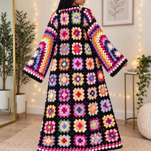 May include: A long, open-front cardigan made from multicolored granny square crochet motifs joined with black yarn. The garment features wide, full-length sleeves and a floor-length hem, showcasing a vibrant pattern of pink, yellow, blue, and purple.