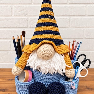 May include: A crochet gnome desk organizer featuring a tall, striped navy and mustard yellow hat, a beige nose, and a white yarn beard. The base is a light blue crochet basket with two side compartments holding pencils, crochet hooks, and scissors.