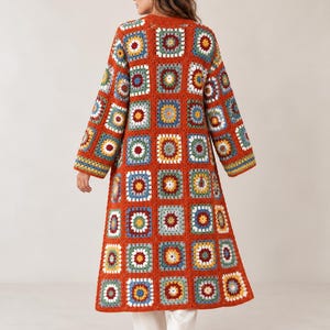 May include: A long, floor-length crochet cardigan constructed from numerous multicolored granny squares. The garment features a dominant burnt orange yarn border connecting each square, with accents of blue, yellow, white, and green.