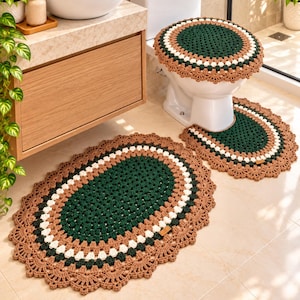 May include: A three-piece crochet bathroom set featuring a dark green, white, and tan color palette. The set includes an oval floor rug, a U-shaped pedestal mat, and a round toilet lid cover, all finished with a decorative scalloped edge.