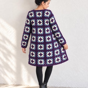 Könnte beinhalten: Ein knielanger Häkelmantel aus quadratischen Motiven in den Farben Dunkelviolett, Hellblau und Creme. Das Kleidungsstück verfügt über lange Ärmel, einen Rundhalsausschnitt und eine durchgehende Knopfleiste mit einem geometrischen Muster.