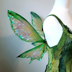 May include: A green fairy costume with iridescent wings. The wings are a vibrant green with a shimmering, translucent quality. The dress is made of layered green fabric, creating a textured, organic look. The costume is displayed on a white mannequin.