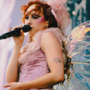 May include: A woman with red hair and fairy wings sings into a microphone. She is wearing a pink dress with sequins and a pink flower crown.