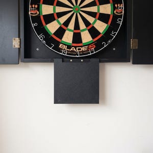 Op de afbeelding: Een zwarte dartbordkast met een Blade 5 dartbord, voorzien van een cirkelvormig doel verdeeld in zwarte, witte, rode en groene segmenten. De kast heeft scharnierende deuren met scoreborden voor Way 01 en Home, en een zwart paneel eronder.