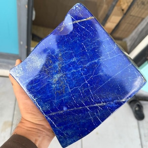 Puede incluir: Una pieza cuadrada y pulida de piedra lapislázuli de color azul intenso, con vetas naturales de calcita blanca e inclusiones de pirita dorada visibles en su superficie lisa y reflectante.
