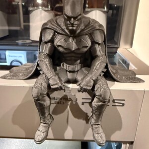 Pode incluir: Uma figura do Batman em preto mate impressa em 3D na posição sentada, com uma máscara detalhada, armadura musculada, uma capa fluida e um emblema de morcego no peito. A figura segura um pequeno acessório em forma de morcego, exibindo as linhas de camada visíveis do processo de impressão.