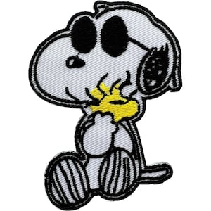 Peut inclure: Un écusson brodé représentant un chien de dessin animé blanc avec des oreilles noires et de grandes lunettes de soleil noires. Le chien est assis et tient un petit oiseau jaune vif dans ses mains. Le motif est bordé de fil noir épais.