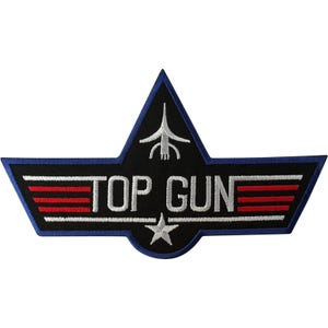May include: A black embroidered patch with a blue border featuring the white text TOP GUN in the centre. The design includes a white stylised aircraft icon above the text, a white star below, and three horizontal red stripes on each side.