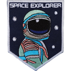 Könnte beinhalten: Ein schildförmiger, bestickter Aufnäher mit dunkelblauem Hintergrund und dem weißen Schriftzug SPACE EXPLORER. Das Motiv zeigt einen Astronauten in einem hellgrünen Anzug mit blauen und orangefarbenen Akzenten, umgeben von kleinen weißen Sternen.