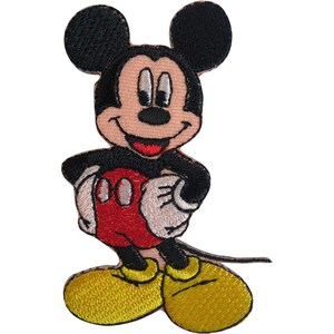 May include: An embroidered fabric patch featuring the character Mickey Mouse. The design shows the character with black ears, a peach-coloured face, red shorts with two white oval buttons, white gloves, and large yellow shoes, all outlined in black thread.