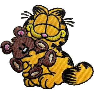 Op de afbeelding: Een geborduurde patch met een gele cartoonkat met zwarte strepen en een brede grijns, die een kleine bruine teddybeer met roze binnenoren vasthoudt. Het ontwerp is omlijnd met zwart garen en toont gedetailleerd stikwerk over het hele figuur.