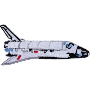 May include: An embroidered patch shaped like a space shuttle in profile. The shuttle is primarily white with black accents on the nose and tail fin, featuring small blue details near the cockpit and red markings on the side of the fuselage.