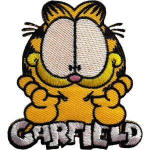 Puede incluir: Un parche bordado que representa al personaje de dibujos animados Garfield, un gato amarillo con rayas negras, grandes ojos blancos y una nariz rosa. El personaje se muestra desde el pecho con las manos cerca de la barbilla. El nombre GARFIELD está en letras blancas.
