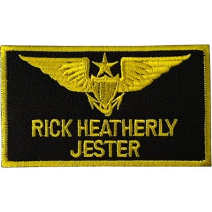 May include: A rectangular black embroidered patch with a yellow border. The design features a yellow embroidered emblem of wings, a star, and an anchor. Below the emblem, the text RICK HEATHERLY JESTER is embroidered in yellow capital letters.