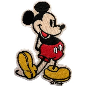 May include: An embroidered iron-on patch featuring the character Mickey Mouse. The design shows the character with black ears, a cream-colored face, a red shirt, red shorts with two white buttons, and large yellow shoes. The patch has a white border.