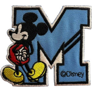 May include: An embroidered patch featuring the letter M in light blue with a thick black border. Mickey Mouse is depicted in black, wearing red shorts and yellow shoes, standing in front of the letter. The text Disney is printed in black.