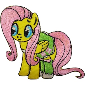 May include: An embroidered patch featuring a yellow pony character with a long, flowing pink mane and tail. The pony has large blue eyes and is wearing a green garment with a small pink pattern. The design is outlined in black thread.