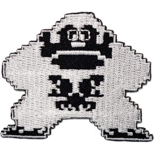 May include: A black and white embroidered patch featuring a pixelated character design. The figure has a blocky, wide-set shape with a distinct head, broad shoulders, and thick legs, rendered in a high-contrast, retro-style digital aesthetic.