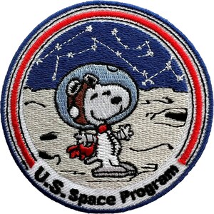 May include: A circular embroidered patch featuring a white cartoon dog wearing a blue space helmet and a red scarf on a grey lunar surface. The background is dark blue with white star constellations. The border is red and blue with the text U.S. Space Program.