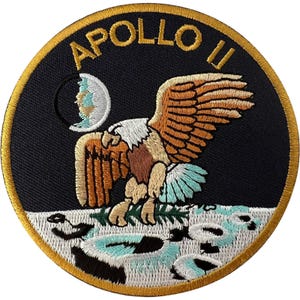 May include: A circular embroidered patch featuring a black background with a gold border. The text APOLLO 11 is stitched in gold at the top. A bald eagle with brown and white feathers descends towards a white lunar surface, holding an olive branch.
