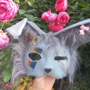Könnte beinhalten: Eine graue Tiermaske mit langen, pelzigen Ohren, einer kleinen schwarzen Nase und netzbespannten Augenöffnungen. Die Maske ist mit einem regenbogenfarbenen Streifen am rechten Auge, einem silbernen Stern am linken Auge, einer Haarspange und einer rosa Schleife verziert.