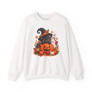 May include: A white long-sleeve sweatshirt featuring a central graphic of a black cat wearing a witch hat, sitting inside a large orange jack-o'-lantern. A small white ghost, autumn leaves, a lantern, and orange stars surround the cat.