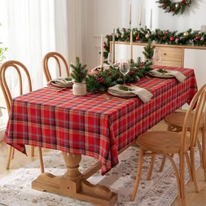 May include: A rectangular dining table covered with a red, yellow, and blue plaid tablecloth. The table is set with white plates, folded napkins, gold cutlery, and a centerpiece of evergreen branches, pinecones, and red berries with candles.