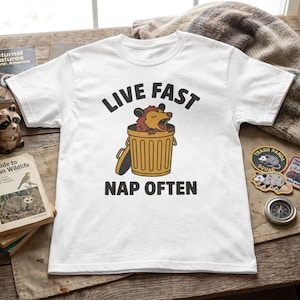 Könnte beinhalten: Ein weißes Kurzarm-T-Shirt mit einer Grafik eines Opossums in einer gelben Mülltonne. Der Text über der Grafik lautet LIVE FAST und der Text darunter NAP OFTEN in fetter schwarzer Schrift, zentriert auf der Vorderseite des Kleidungsstücks.