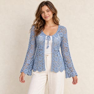 Könnte beinhalten: Eine hellblaue Häkelstrickjacke mit Blumenmuster, langen ausgestellten Ärmeln und einem gewellten Saum. Das Kleidungsstück hat ein offenes Design mit einem dünnen Bindeband am Ausschnitt, getragen über einem weißen Oberteil und einer hellen Hose.