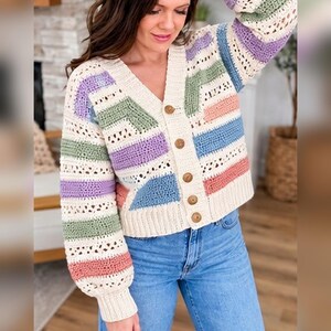 Könnte beinhalten: Eine cremefarbene Häkelstrickjacke mit V-Ausschnitt, langen Ärmeln und einer Knopfleiste mit vier braunen Knöpfen. Das Kleidungsstück ist mit horizontalen Streifen in den Farben Lavendel, Salbeigrün, Blau und Korallenrosa verziert.