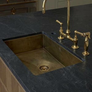 May include: A rectangular brass kitchen sink set into a dark gray stone countertop. The sink features a matching brass drain and is paired with a tall, arched brass faucet with two side handles and a separate brass side sprayer attachment.