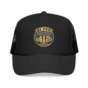 May include: A black trucker hat featuring a circular graphic on the front. The design includes the text YINZER in gold, a bridge illustration, the number 412 in large gold digits, and the text PITTSBURGH, PA below, all enclosed in a gold circular border.